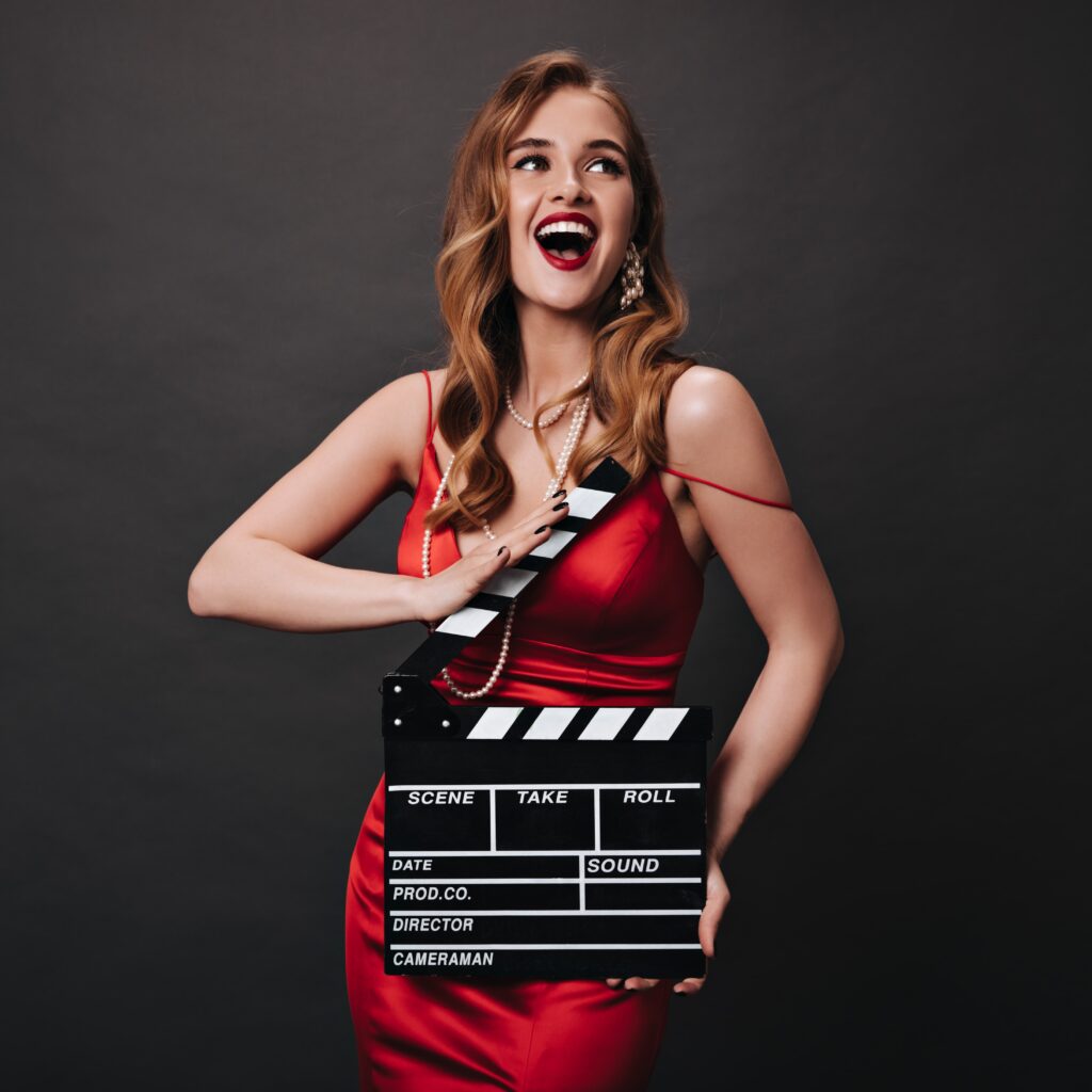 young-lady-with-wavy-hair-beautiful-makeup-posing-with-clapperboard-happy-emotional-woman-red-festive-dress-smiling-black-background