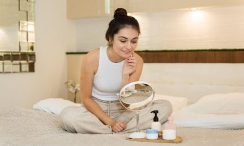  young-caucasian-woman-is-sitting-on the-bed-and-looking-in-the-mirror-skin-care-products-nearby